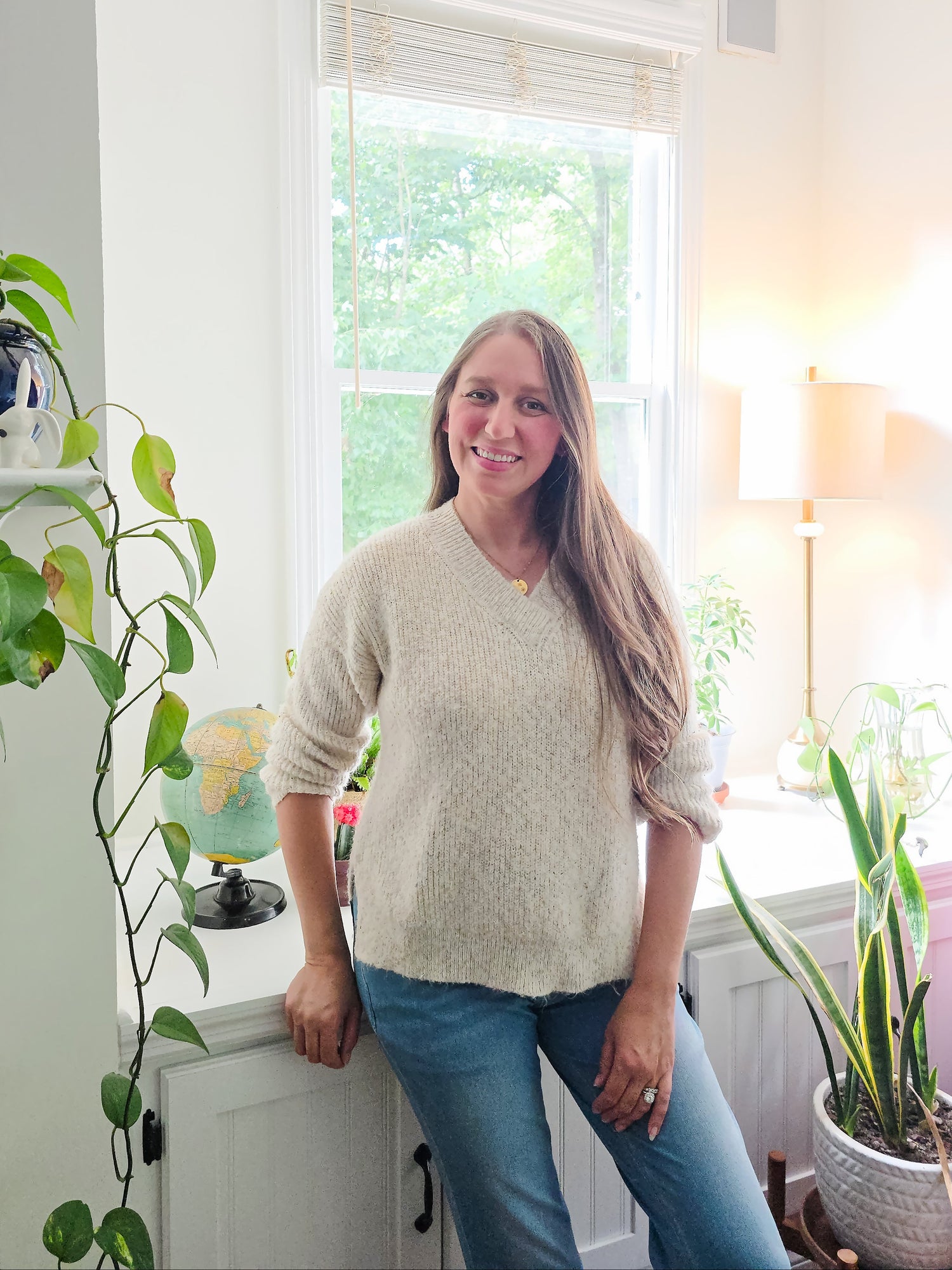 Kelly Zayas Bass stands in a bright room wearing a cream-colored knit sweater and blue jeans, leaning against a white cabinet next to a window and houseplants.